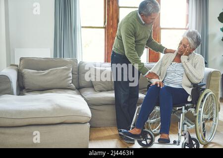 Nous y partons ensemble. Prise de vue d'une femme âgée inquiète assise dans un fauteuil roulant tout en étant soutenue et tenue par son mari à l'intérieur Banque D'Images