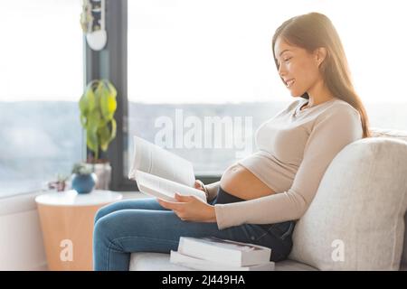 Femme asiatique enceinte lisant des livres prénatals se préparant au travail, à la naissance, au nouveau-né, assis sur un canapé à la maison. Mode de vie pendant la grossesse Banque D'Images