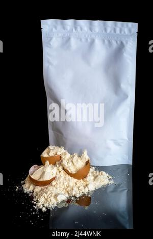 Tas de poudre de protéine d'œuf et coquilles d'œuf, maquette de paquet blanc avec espace de copie, isolé sur fond noir avec réflexion miroir sur la table. Sport Banque D'Images