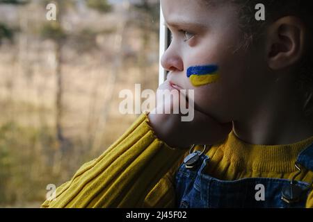 Gros plan d'une petite fille triste avec le drapeau de l'Ukraine sur le visage assis seul dans le seuil de fenêtre à la maison. Enfant malheureux sur fond de nature. La paix, pas de guerre, il Banque D'Images