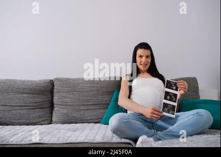 Au cours d'une grossesse avancée, une femme blanche noire aux poils longs s'appuie sur des oreillers verts.une femme enceinte au ventre énorme montre une image échographique des bébés. Banque D'Images