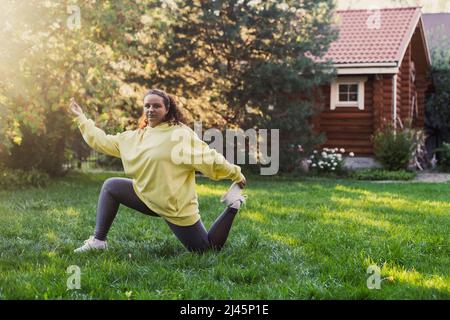 Femme d'âge moyen surpondérée faisant du yoga dans des vêtements pour le sport sur l'herbe verte dans la cour avec maison de campagne en bois et de grands arbres en arrière-plan Banque D'Images