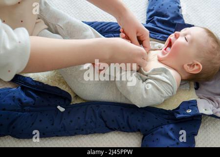 Une mère femme met des vêtements chauds pour un bébé garçon. Maman habille un enfant heureux dans des vêtements d'hiver sur le lit. Banque D'Images