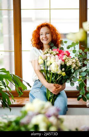 Belle fleuriste femme avec des cheveux rouges bouclés tenant des fleurs fraîchement fait bouquet de fleurs avec des pivoines, roses, eucalyptus sur fond de fenêtre Banque D'Images