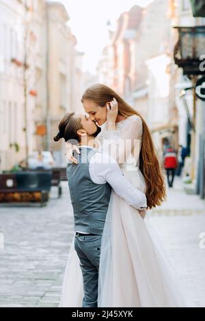 Beau marié tient sa mariée dans les mains embrassant sur les lèvres sur fond de rue du matin Banque D'Images