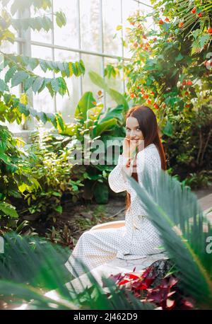 Fille fragile avec de longs cheveux rouges assis dans une serre de plantes tropicales. Banque D'Images