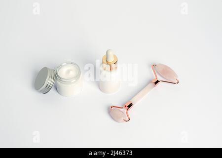Crème dans un pot, bouteille de sérum et rouleau facial en quartz rose cristal sur fond blanc, vue du dessus. Banque D'Images