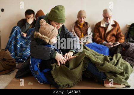 Vue latérale de la jeune mère tenant son fils endormi tout en se cachant dans un refuge pour réfugiés et assise sur le sol recouvert de couvertures Banque D'Images