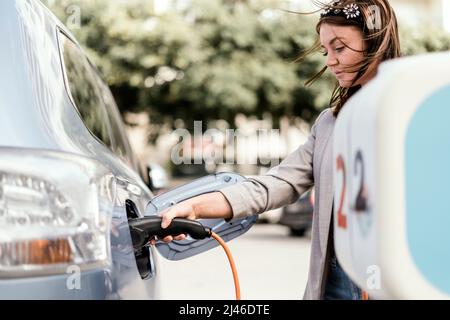 Une jeune femme charge une voiture électrique en milieu urbain Banque D'Images