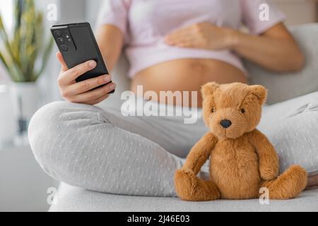 Application grossesse pour cours en ligne sur la maternité prénatale. Femme enceinte utilisant un téléphone portable avec l'accent sur bébé jouet ours en peluche. Pépinière, nouveau-né Banque D'Images