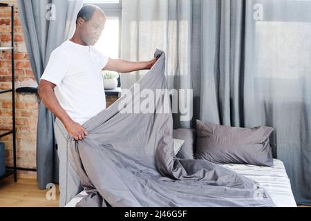 Un vieil homme afro-américain contemporain en t-shirt faisant un lit avec couverture grise le matin après le sommeil et les exercices physiques Banque D'Images