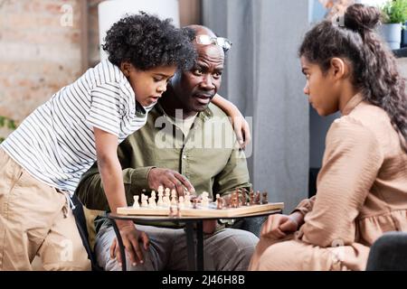 Un homme afro-américain senior et ses deux petits-enfants jouant aux échecs ensemble tout en étant assis à une petite table avec des figures à bord Banque D'Images