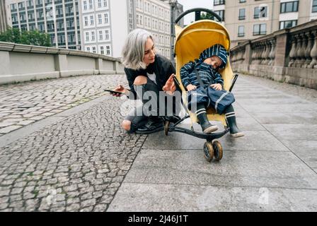 La mère a piedé dans la poussette avec son jeune fils assis sur le trottoir Banque D'Images