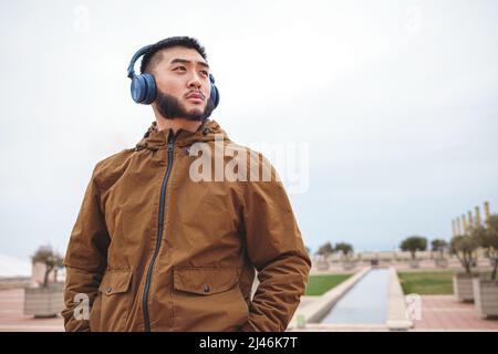 Homme asiatique sérieux en vêtements d'extérieur à l'écoute de la musique dans le parc Banque D'Images