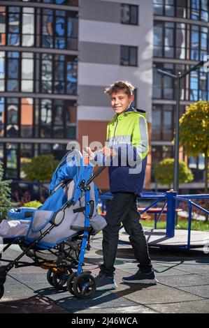 Portrait d'un jeune garçon qui s'occupe de passer du temps avec un frère nouveau-né à l'aire de jeux. Jeune adolescent marchant avec bébé dans une poussette dans la rue dans la cour moderne des immeubles résidentiels de la ville. Banque D'Images