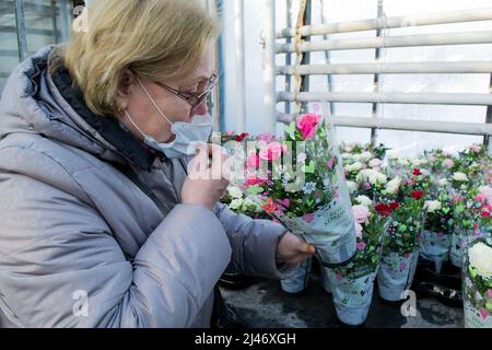 Moscou, Russie, février 2022: Femme âgée dans un masque médical sniffs roses dans des pots et enveloppé: Vente dans la serre. Fleurs en cadeau. Banque D'Images