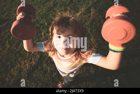 Sport amusant pour enfants. Enfant avec haltères dans le parc. Petit garçon fort. Visage d'enfant amusant. Développement des enfants et exercice sain et fort. Grand angle. Banque D'Images