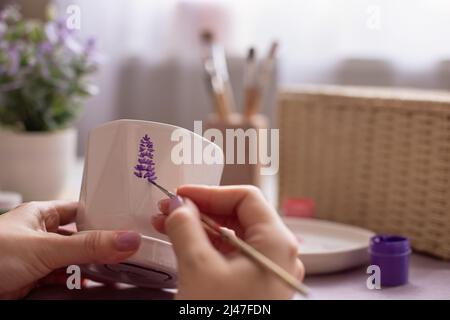 Gros plan des mains de femmes peignent avec un pinceau une branche d'une lavande pourpre , sur un pot en céramique blanche. Banque D'Images