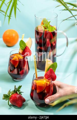 Main féminine tenant un verre avec un cocktail traditionnel espagnol Sangria décoré avec des morceaux de fraise fraîche, pincée de menthe et de paille de papier écologique. Banque D'Images