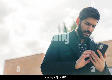 Beau barbu latino-américain dans des vêtements noirs regardant dans le téléphone confus moyen de gros plan extérieur urbain fond. Photo de haute qualité Banque D'Images