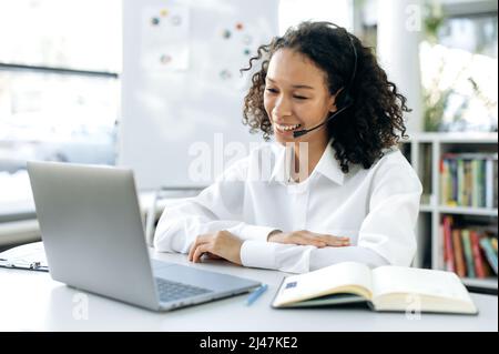 Jeune femme afro-américaine amicale, opérateur de hotline, employé de service d'assistance, consultant en ligne, assis à un écran d'ordinateur portable, conduit une consultation vidéo avec un client, écoute attentivement, sourit Banque D'Images