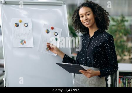 Jeune femme afro-américaine réussie et déterminée, vêtue de vêtements formels, gestionnaire financier, économiste, Trader, se tient dans un bureau près d'un tableau blanc, présente son rapport de projet, souriant amical Banque D'Images