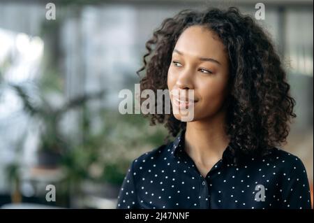 Gros plan d'une jeune femme afro-américaine élégante et attirante avec cheveux bouclés, gestionnaire, commerçant, SPÉCIALISTE INFORMATIQUE, debout dans un bureau moderne, regardant sur le côté, pensant, rêvant, souriant Banque D'Images