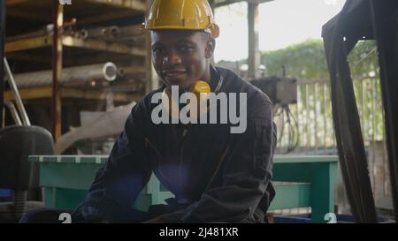 Portrait américain industriel noir jeune homme de travail souriant avec casque jaune devant la machine, heureux ingénieur assis au travail dans l'industrie fac Banque D'Images