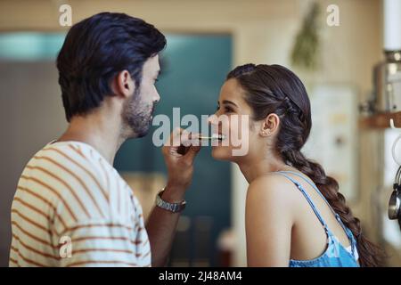 L'amour rend tout le goût meilleur. Photo d'un jeune couple affectueux qui se grignote tout en préparant un repas dans sa cuisine. Banque D'Images