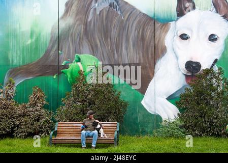 La fresque de l'artiste de rue « Sugar » à porter Street dans le centre-ville de Prahran, Melbourne, domine un petit chien et son maître est assis sur un banc de parc. Banque D'Images