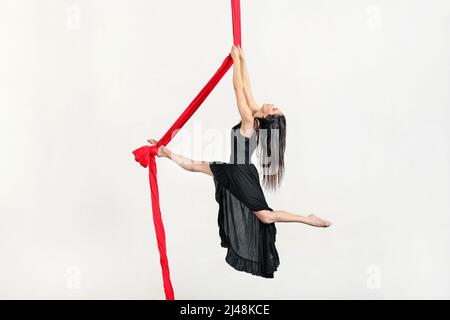 Vue latérale du corps de la femme active aerialist en robe faisant un tour sur les silks aériens rouges sur fond blanc en studio Banque D'Images