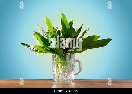 beau bouquet de lys frais de la vallée dans une tasse en verre sur fond bleu de table en bois Banque D'Images
