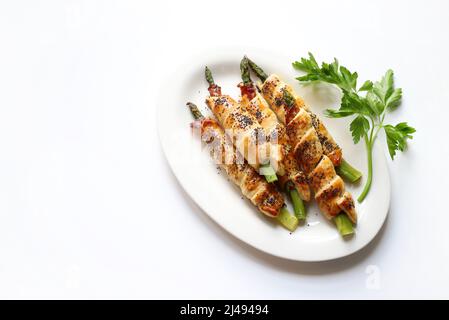 Asperges fraîchement cuites au four enveloppées de prosciutto et de pâte feuilletée, décorées de graines de pavot sur fond blanc. Saison de printemps. Directement au-dessus. Banque D'Images