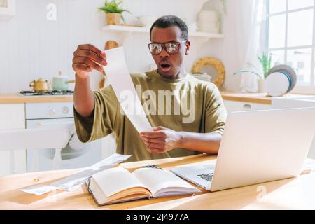 L'homme afro-américain regarde les chèques en comptant la quantité d'argent dépensé dans le shopping. Guy en lunettes se met en colère assis dans la cuisine à la maison vue de près Banque D'Images