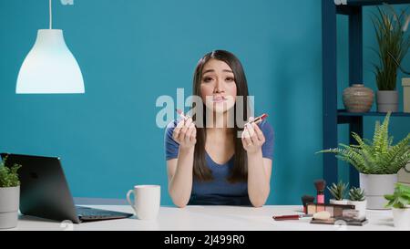 POV de l'influenceur asiatique faisant la recommandation de rouge à lèvres en studio, le tournage de la révision de maquillage avec des produits cosmétiques sur l'appareil photo. Une femme blogueuse enregistre des vidéos sur les réseaux sociaux. Cliché sur trépied. Banque D'Images