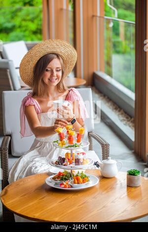 Femme en robe blanche et chapeau assis sur une chaise au café moderne avec vue sur la mer Banque D'Images