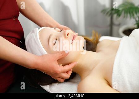 Calme jolie femme avec les yeux fermés couché sur le corps de la table de massage recouvert d'une serviette blanche subissant un massage du visage dans le salon de spa. Banque D'Images