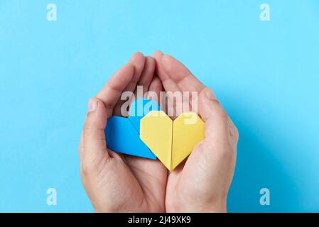 Mains d'une personne méconnaissable tenant et protégeant une paire de coeurs d'origami bleu et jaune, couleurs du drapeau ukrainien. Image symbolique, solide Banque D'Images