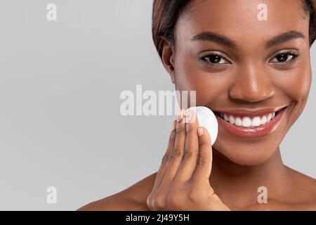 Souriante jolie femme afro-américaine millénaire essuyant son visage avec un tampon en coton, isolé sur fond gris, gros plan Banque D'Images