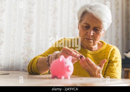 Investissement et épargne. Pauvre caucasienne retraité gris-cheveux dame en chandail jaune regarde son argent et met des pièces à sa banque de porc rose. Photo de haute qualité Banque D'Images