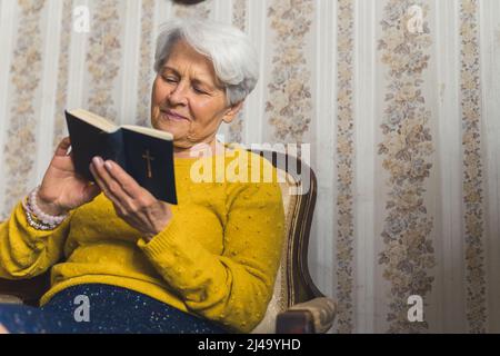 Grand-mère caucasienne âgée de 70s ans, assise sur une chaise à l'ancienne et listant pacifiquement la Bible. La spiritualité dans le concept de la vieillesse. . Photo de haute qualité Banque D'Images