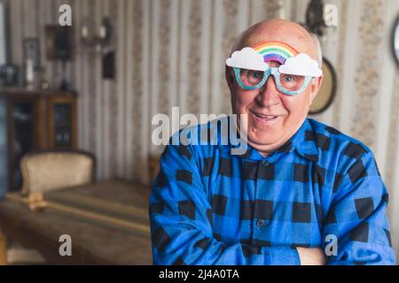 Un vieil homme chauve européen dans une chemise en flanelle bleu et des lunettes arc-en-ciel regardant la caméra et se tenant dans son salon d'époque. Photo de haute qualité Banque D'Images