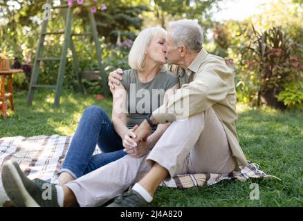 Couple senior marié se reposant à l'extérieur et embrassant, appréciant des vacances tout en étant assis sur une couverture sur l'herbe dans le jardin Banque D'Images