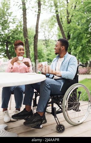 Jeune Afro-américain en fauteuil roulant ayant rendez-vous avec une belle femme au café en plein air, en appréciant le café au parc Banque D'Images