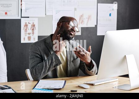 Un médecin africain de sexe masculin touche sa gorge et parle au patient en ligne sur un ordinateur assis sur le lieu de travail au bureau Banque D'Images