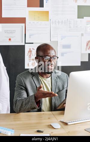 Un homme africain médecin en lunettes assis à une table devant un moniteur d'ordinateur et ayant une consultation en ligne au bureau Banque D'Images