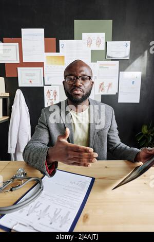 Spécialiste médical africain de lunettes utilisant des cartes et des documents médicaux tout en parlant pendant la conférence en ligne sur ordinateur au bureau Banque D'Images