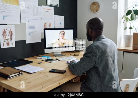 Vue arrière du généraliste africain assis devant le moniteur d'ordinateur et ayant une réunion en ligne avec son patient Banque D'Images