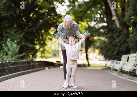 Mère et fille musulmanes positives avec des cloches dans le parc d'été. Femme arabe sportive montrant son petit enfant s'ébaissant avec des haltères, se relève Banque D'Images