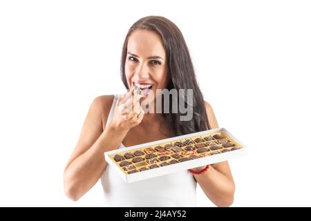 Une bonne femme aux cheveux foncés prend une bouchée d'un bonbon tout en tenant une boîte de bonbons pleine. Arrière-plan blanc isolé. Banque D'Images